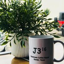 A white J316 Downtown Coffee House mug beside a potted plant.
